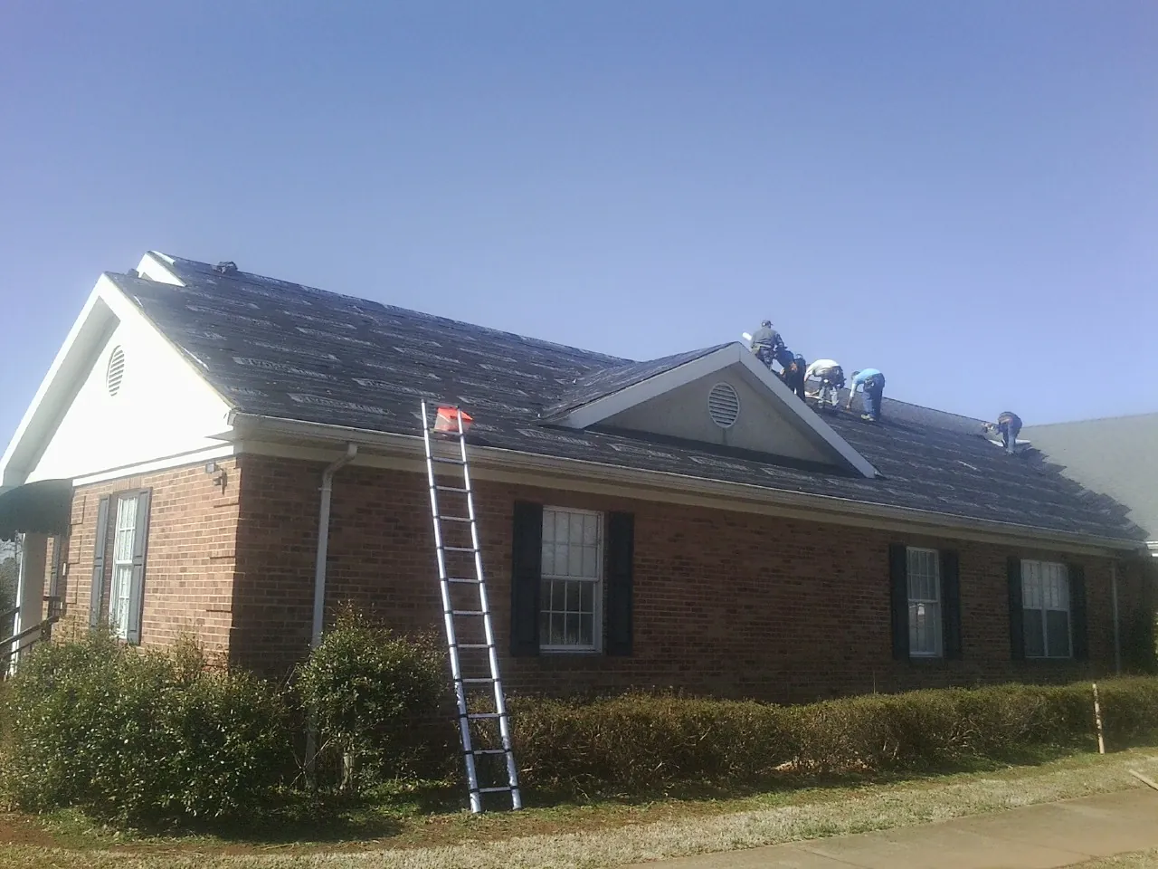 Crew tearing shingles off a brick ranch roof with a ladder set — Emergency Roof Repair work by our crew in Penrose